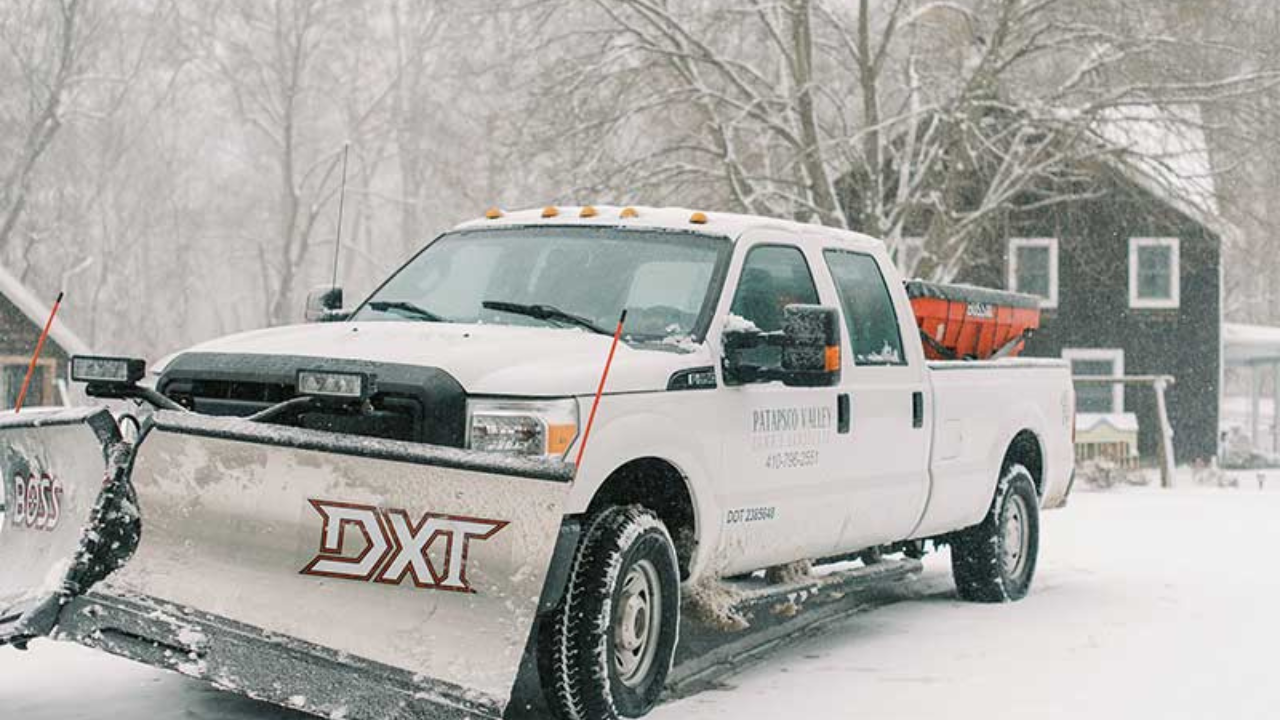 A Patapsco Valley Lawn & Landscape Pickup Truck equipped with a snow plow while it is snowing